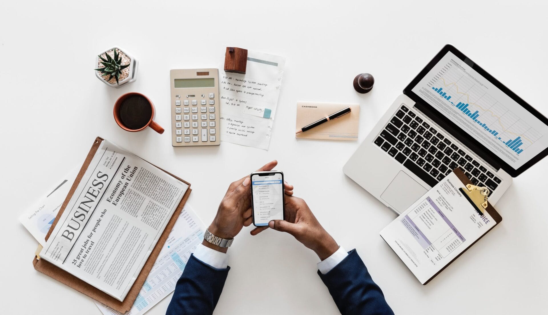 Business person looking at phone on a desk with newspaper and laptop.