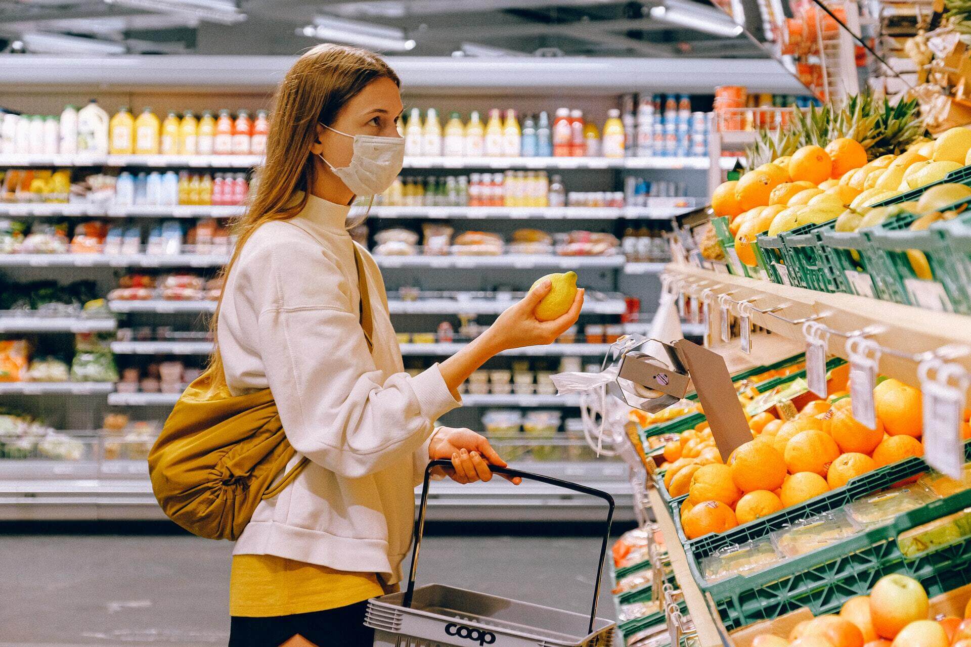 A women in a yellow t-shirt and beige jacket holding a fruit at the grocery store